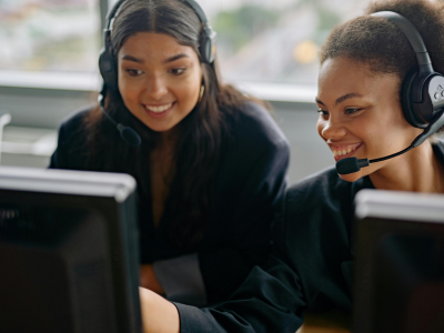 Zwei Mitarbeiterinnen tragen Headsets und sitzen vor Computerbildschirmen.